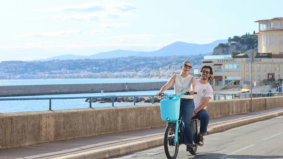 男性と、その同伴者でサングラスをかけた女性が、青い湾に沿った海辺の遊歩道を一台の自転車で進んでいます。二人とも笑顔です。